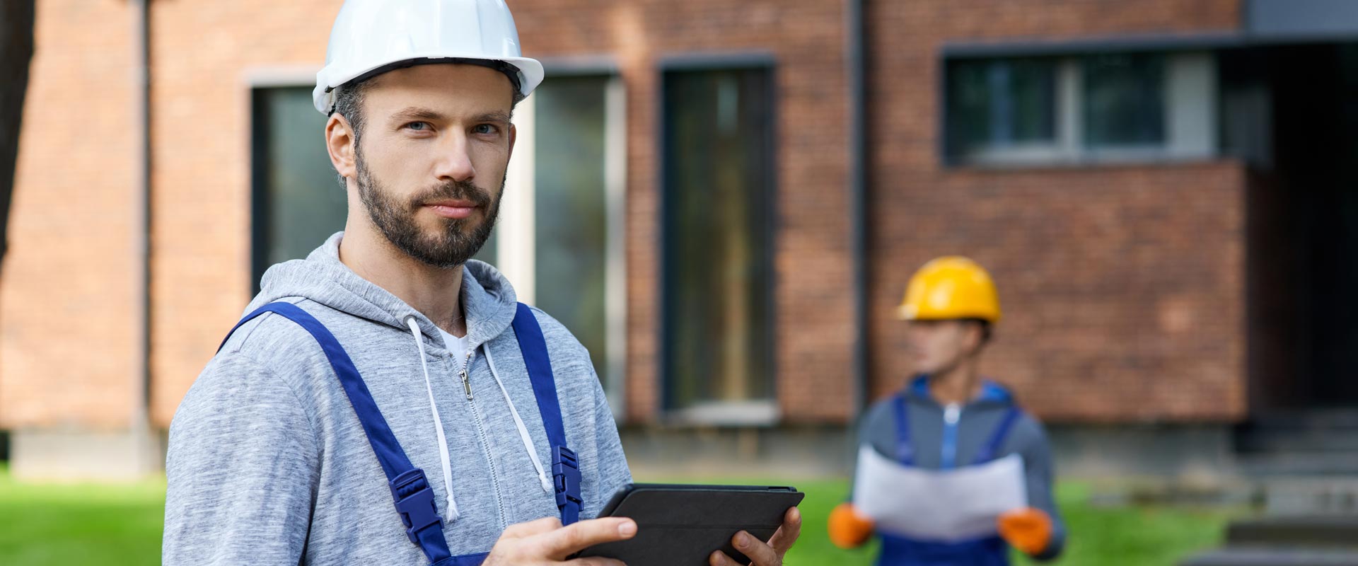 Construction worker in a white hard hat holds a tablet, with a colleague in a yellow helmet
