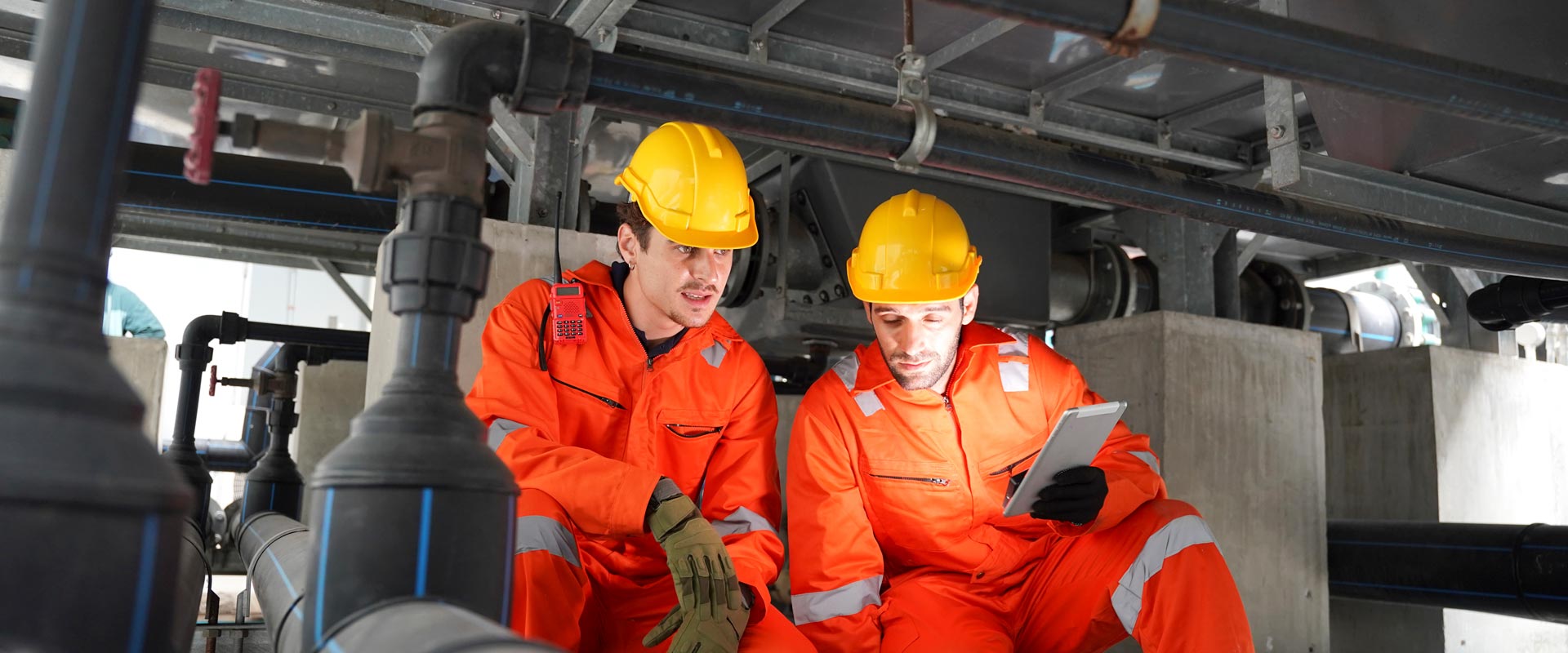 Two workers in orange uniforms and yellow helmets inspect pipes in an industrial setting