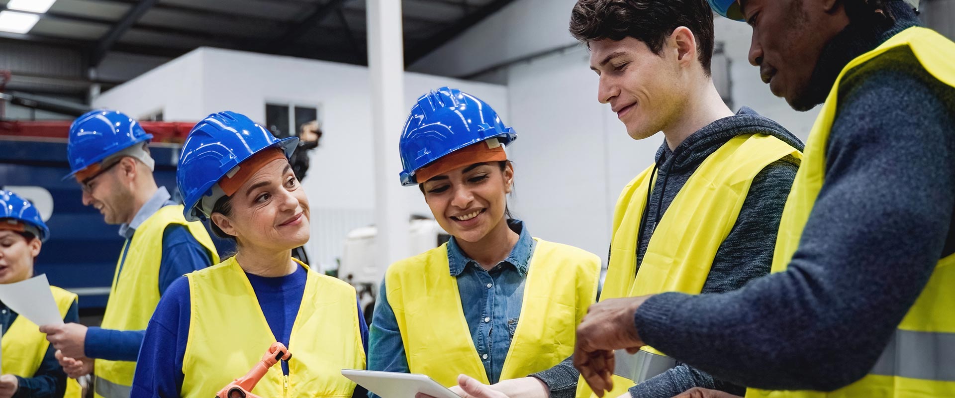 Workers in blue helmets and yellow vests discuss plans in a warehouse
