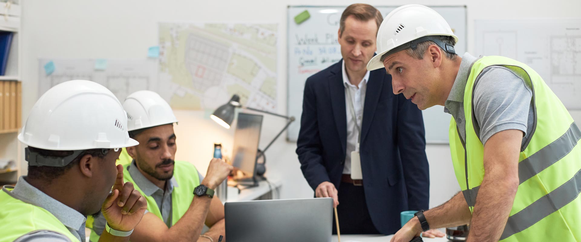 Engineers in hard hats and vests discuss plans at a table, while a manager in a suit observes