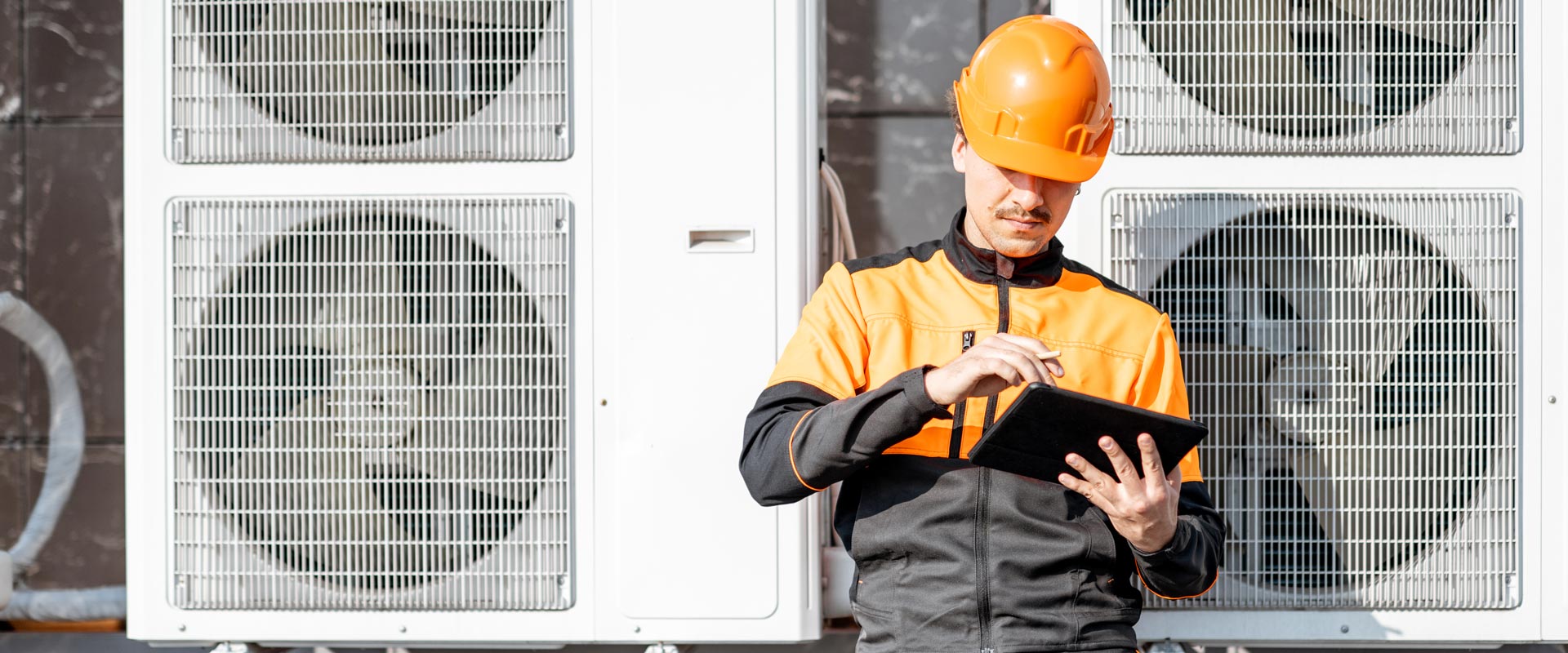 Worker in orange safety gear uses a tablet in front of large industrial air conditioning units