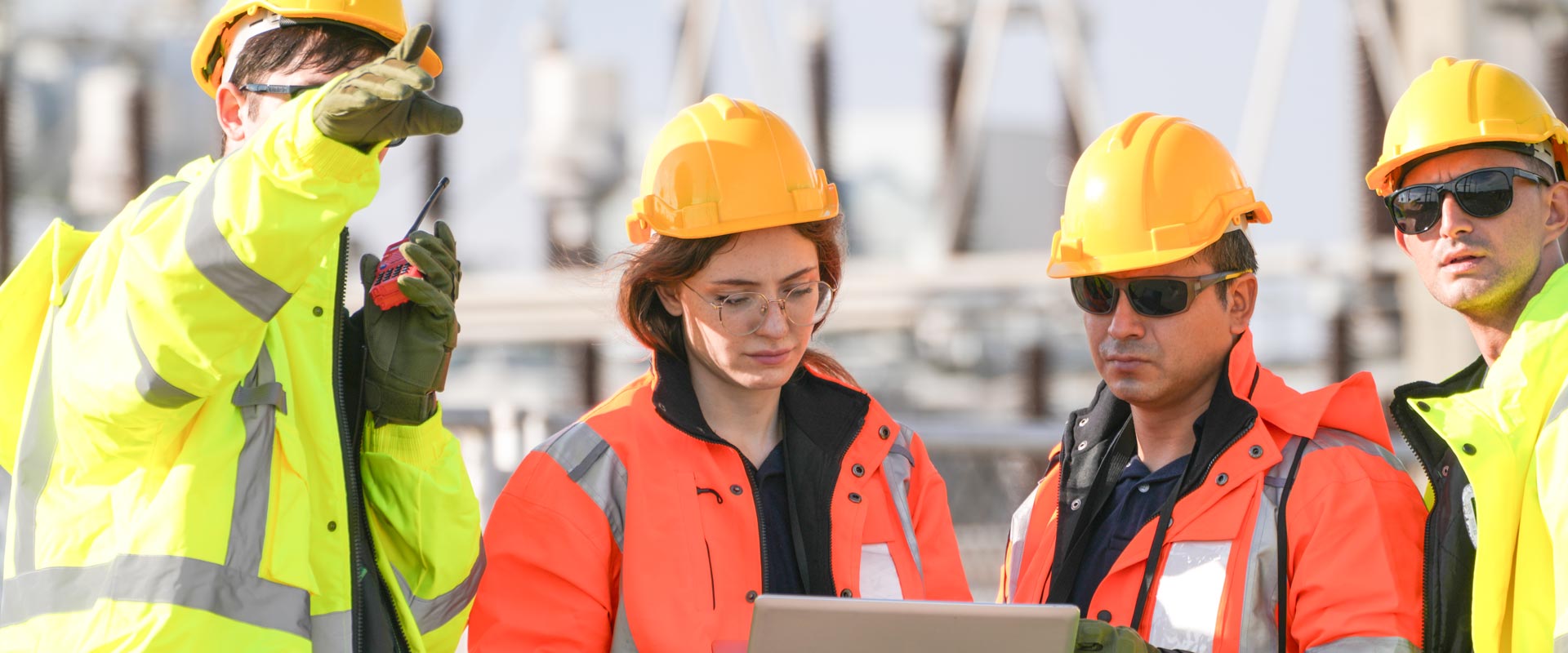 Engineers in hard hats and safety jackets collaborate outdoors, reviewing plans on a laptop at a construction site
