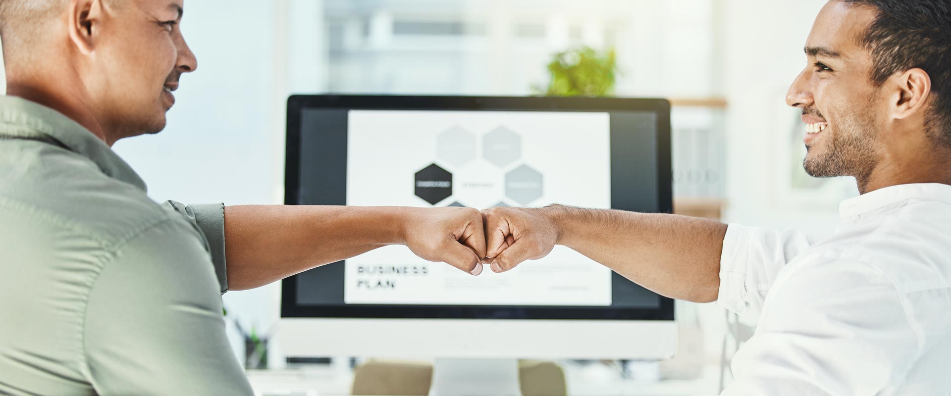 Two men smiling, fist-bumping in an office with a computer screen displaying a business plan in the background