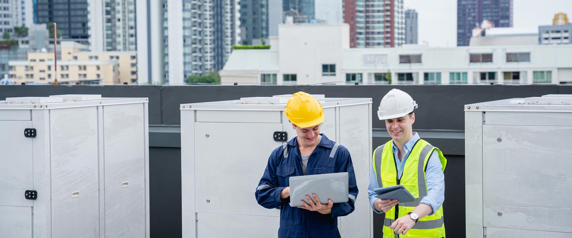 Two engineers in hard hats and vests discuss work on a rooftop, using a laptop and tablet, with city buildings in the background