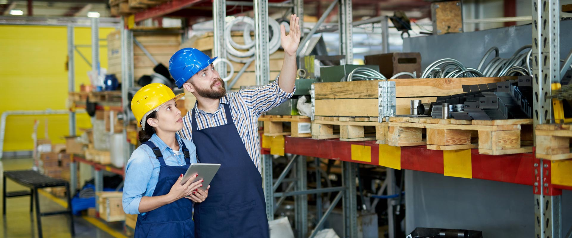 Two workers in hard hats and aprons discuss inventory in a warehouse, one holding a tablet, surrounded by shelves and pallets